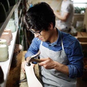 Young man in apron sitting by his workplace and hammering nails into sole of footwear workpiece